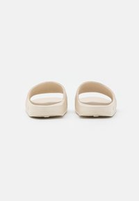 Sandali beige con un design minimalista, caratterizzati da un'apertura frontale ampia, una texture liscia, una forma bassa e senza hardware visibile.