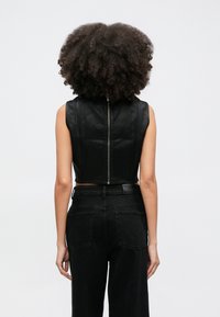 Žena s kučeravými vlasmi má na sebe čierny kožený crop top bez rukávov so zipsom na chrbte a čierne vysokopridržané džínsy, stojaca na bielom pozadí.