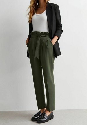 Femme portant un pantalon à taille paperbag vert olive avec ceinture, débardeur blanc, blazer noir et mocassins noirs, debout devant un fond blanc.