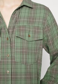 Chemise à carreaux verte et marron à boutons, avec poche poitrine et col, portée par une personne dont le cou et le bras sont partiellement visibles.