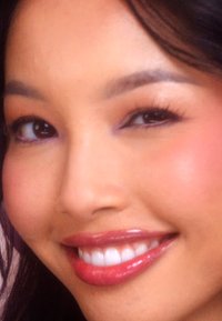 Close-up van een glimlachend gezicht met gladde, lichte huid, glanzende lippen in een roze tint, en donkere, lange wimpers onder zacht gebogen wenkbrauwen.