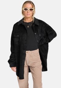 Schwarze Cordjacke mit vorderen Knöpfen und zwei Brusttaschen, kombiniert mit einem schwarzen Oberteil und beigen Cordhosen.