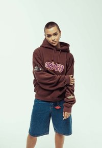 Sudadera marrón con el gráfico "Grimy" en púrpura, con diseños laterales y un ajuste holgado, combinada con pantalones cortos de mezclilla azules y un detalle de colgante. 