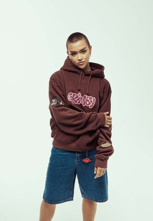 Sudadera marrón con el gráfico "Grimy" en púrpura, con diseños laterales y un ajuste holgado, combinada con pantalones cortos de mezclilla azules y un detalle de colgante. 