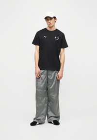 Νέος άνδρας που φοράει λευκό καπέλο, μαύρο t-shirt Puma BMW Motorsport, γυαλιστερό ασημί φαρδύ παντελόνι και μαύρα και λευκά παπούτσια, μπροστά σε απλό λευκό φόντο.