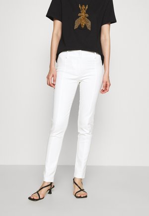 Frau trägt weiße Slim-Fit-Hosen, schwarze Riemchensandalen und ein schwarzes T-Shirt mit einem großen goldenen Insekten-Design auf der Brust.