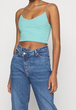 Persona delgada con un top corto de canalé turquesa con tirantes finos y vaqueros azules de talle alto y sueltos con la cintura desabrochada.