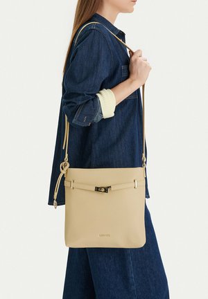 Mujer con conjunto de mezclilla que lleva un bolso cruzado de cuero beige con broche dorado y correa ajustable sobre el hombro.