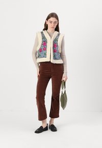 Faux-Fur-Weste mit mehrfarbigem Blumenstickereien, kombiniert mit braunen Cordhosen und schwarzen Slippern. Model hält eine grüne Handtasche.