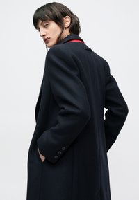 Lange marineblauwe jas met een gestructureerde stof. Voorzien van een geknotte kraag, rode accenten bij de hals en manchetten met knopen. Zakjes aan beide zijden.