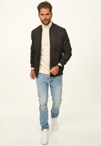 Schwarze Wildleder-Bomberjacke mit hohem Kragen, getragen über einem cremefarbenen Rollkragenpullover, kombiniert mit hellblauen Jeans und weißen Sneakern.