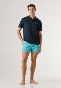 Ung mand stående barfodet iført en mørkeblå polo og turkise badeshorts med fuglemønster mod en ensfarvet baggrund.