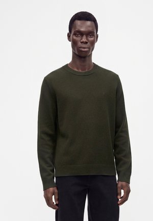 Homme portant un pull vert foncé à manches longues et un pantalon noir, debout devant un fond blanc uni.
