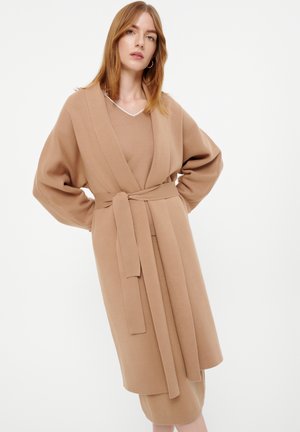 Knielange, wikkelstijl cardigan van zacht, textuurachtig beige stof met een strikriem en brede mouwen. Gelaagd over een bijpassende outfit.