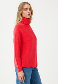 Czerwony sweter z wysokim kołnierzem, wykonany z miękkiego, teksturowanego materiału, z prążkowanymi mankietami i dołem. Połączony z niebieskimi dżinsami dla casualowego wyglądu.