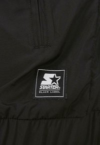 Chaqueta negra con un tejido texturizado y un pequeño parche de logo "Starter Black Label" en blanco. El diseño incluye un detalle de costura tipo pespunte.