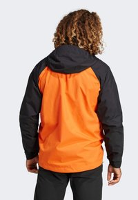 Orange og sort vandafvisende jakke med hætte, der har et strømlinet design, glat tekstur og kontrasterende farveblokering på ærmerne.
