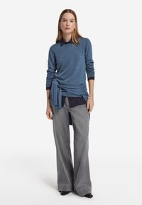 Kvinna stående iklädd en blå tröja knuten i midjan över en mörk skjorta, med vida grå jeans och svarta spetsiga skor.