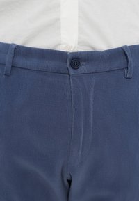Pantalon en velours côtelé bleu marine à la texture lisse, doté d'une fermeture à boutons et de passants pour ceinture à la taille, présenté sur une chemise claire.