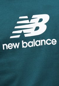 Λευκό λογότυπο και κείμενο New Balance τυπωμένα σε πετρόλ ύφασμα, που δείχνουν το στυλιζαρισμένο "NB" με οριζόντιες γραμμές ταχύτητας και το "new balance" από κάτω.