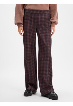 La personne porte un pantalon large à carreaux marron, des chaussures noires épaisses et un haut mauve à manches longues avec des manches bouffantes.