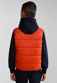 Gilet imbottito rosso con sezioni a motivo trapuntato orizzontali, dotato di cappuccio e indossato sopra una maglia a maniche lunghe blu scuro. Tessuto liscio.