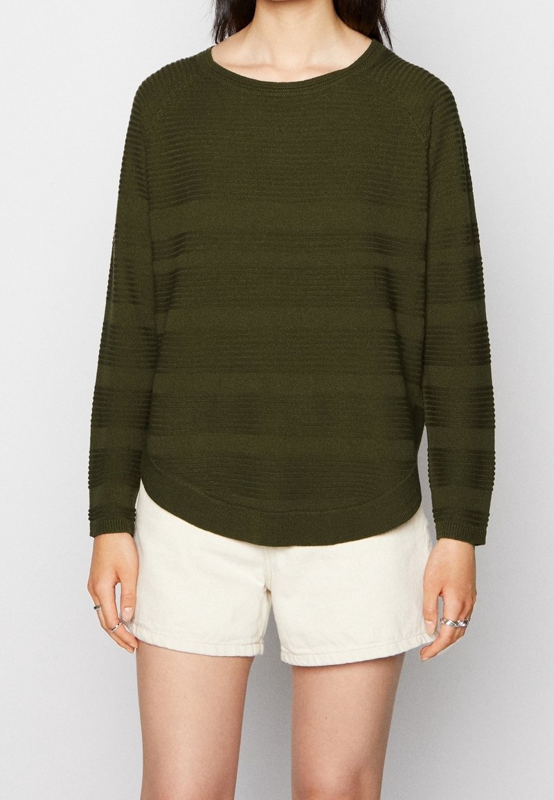 Personne portant un pull côtelé vert foncé à manches longues et un short écru, debout les mains détendues le long du corps.