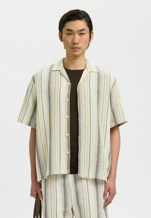 Homme portant une chemise à manches courtes à rayures verticales beige, blanche et marron, et un short assorti, tenant un sac vert olive.