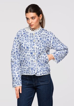 Kvinde iført en hvid jakke med blå blomsterprint og knapper, kombineret med mørkeblå jeans, poserer med den ene hånd på hoften.