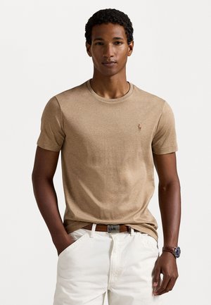 Beige t-skjorte med korte ermer i et mykt stoff, med rund hals og en liten logo på brystet. Satt sammen med hvite bukser.