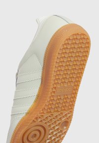 Vita lädersneakers med tre diagonala ränder, en texturerad gummisula i ljus orange och ett mönstrat grepp. Logotypen är präglad på sulan.