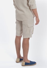 Pantaloni cargo in lino beige con due tasche laterali, orli arrotolati e vestibilità dritta, abbinati a espadrillas blu e beige.