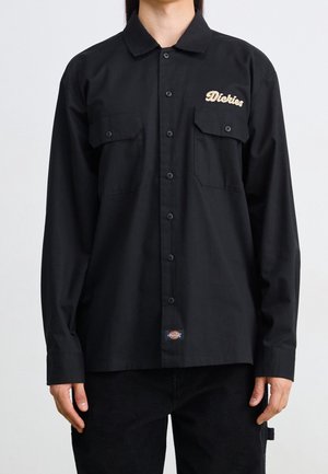 Henkilö, jolla on päällään musta pitkähihainen napillinen paita, jossa on kaksi läppätaskua ja rinnassa brodeerattu "Dickies"-logo.