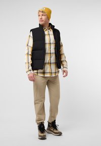 Schwarze gepolsterte Weste über einem gelb-weißen karierten Hemd, beige Hose und schwarz-braunen gemusterten Stiefeln. Gelbe Strickmütze mit Logo.