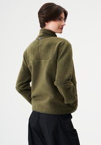 Olivgrüne Fleecejacke aus Kunstfell mit hohem Kragen und einem Logoakzent auf der Rückseite. Verfügt über eine lockere Passform und weiches, strukturiertes Material.