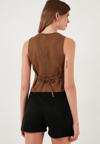 Barna velúr ujjatlan felső, illeszkedő fazonnal és hátkötő részlettel, fekete rövidnadrággal párosítva. Simaság jellemzi, elegáns kialakítás.