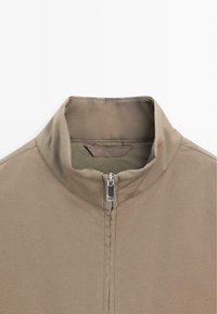 Brun lynlåstrøje med høj krave, letvægtsstof og ren, minimalistisk design med synlige syningsdetaljer.