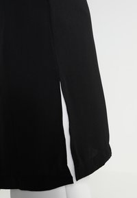 Schwarzes Kleid mit glatter Textur, das einen seitlichen Schlitz und einen geraden Saum aufweist. Der Stoff wirkt leicht und fällt sanft.