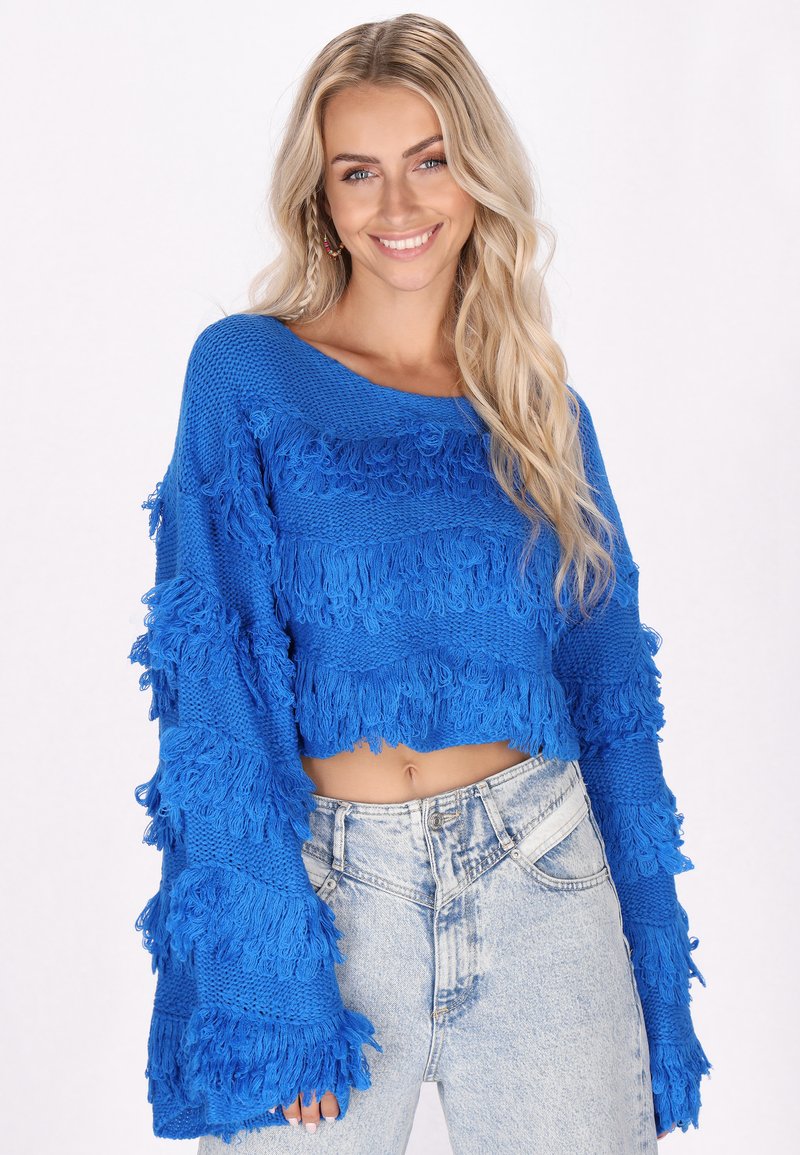 Blauer, kurzer Strickpullover mit strukturierten Fransen, langen Ärmeln und weitem Rundhalsausschnitt. Getragen mit hellblauen, high-waisted Jeans.