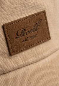 Gorra de tela color beige con un parche de cuero marrón que tiene el texto grabado "Reell est. 1997". La textura es suave y tiene costuras visibles.