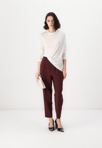 White draped top with long sleeves, paired with burgundy tailored trousers. Model holding a beige clutch and wearing black high-heeled shoes.