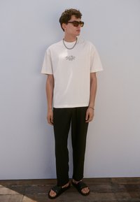 T-shirt color crema con una piccola grafica, pantaloni neri a righe sottili, sandali neri a ciabatta e occhiali da sole. Sfondo neutro.