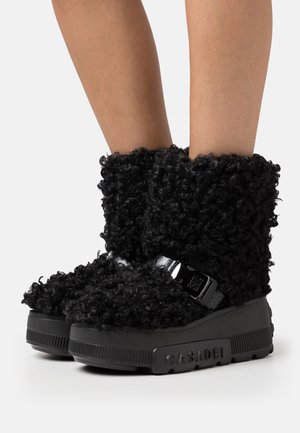 Bottes courtes noires en matière pelucheuse avec un extérieur texturé, dotées d'une bride brillante et d'une semelle noire épaisse. Nom de la marque "CASADEI" embossé sur le côté.