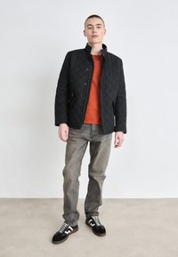 Μαύρο πουκαμισάκι με επένδυση και ψηλό γιακά, φορεμένο πάνω από πορτοκαλί πουκάμισο. Γκρι τζιν με ίσιες γραμμές και μαύρα αθλητικά παπούτσια με λευκές λεπτομέρειες ολοκληρώνουν την εμφάνιση.