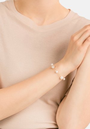 Bracelet en or délicat avec cinq perles blanches rondes et un accent en cristal rond, porté sur un haut beige discret.