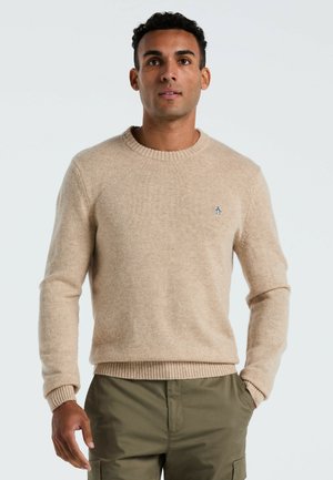 Beige gebreide trui met een ronde halslijn, geribde manchetten en zoom; heeft een klein geborduurd logo op de borst. Draagt over groene broek.