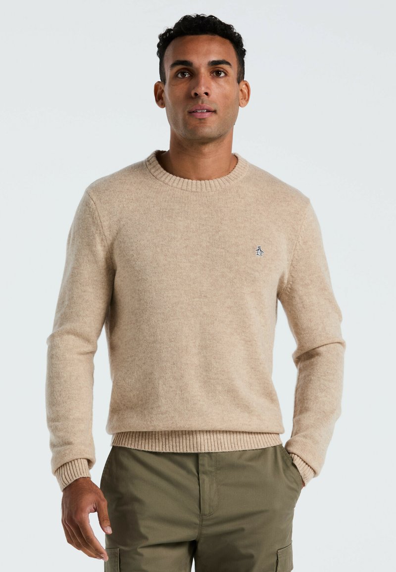 Beige gebreide trui met een ronde halslijn, geribde manchetten en zoom; heeft een klein geborduurd logo op de borst. Draagt over groene broek.