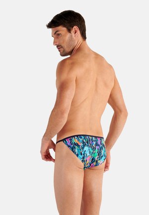 Man with short dark hair wearing colorful abstract-patterned swim briefs, facing backward and looking over his shoulder against a plain background.