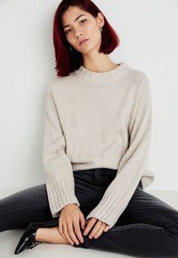 Suéter de punto beige con puños acanalados y cuello redondo, combinado con jeans oscuros y zapatos negros de punta. Diseño sencillo y de corte relajado.