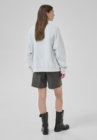 Sweatshirt oversize blu chiaro con polsini a costine, abbinato a pantaloni corti grigi sartoriali. Il modello indossa stivali neri al ginocchio con fibbie.
